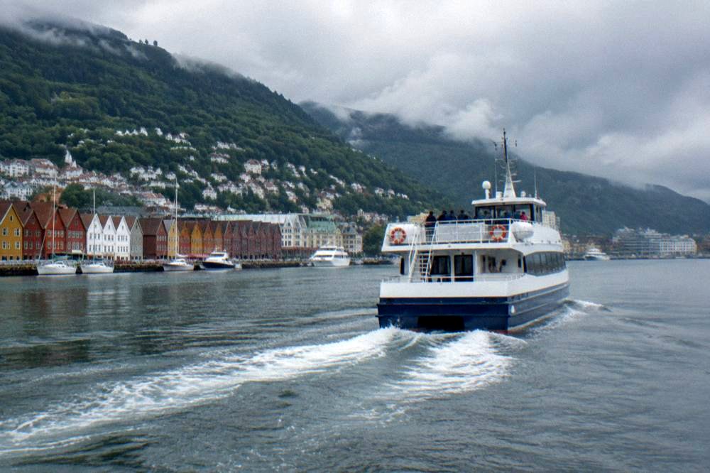 A tour boat leaving Bergen harbour with forested hills rising behind the city