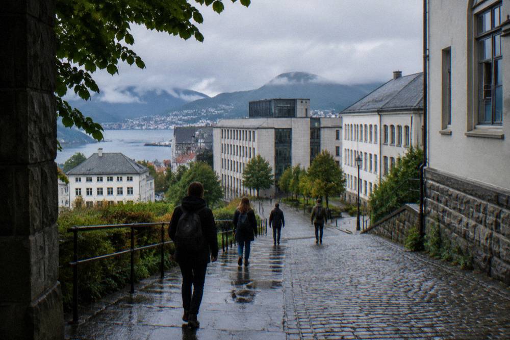 People walking up a sloping stone street in Bergen toward a cluster of university buildings under overcast skies.