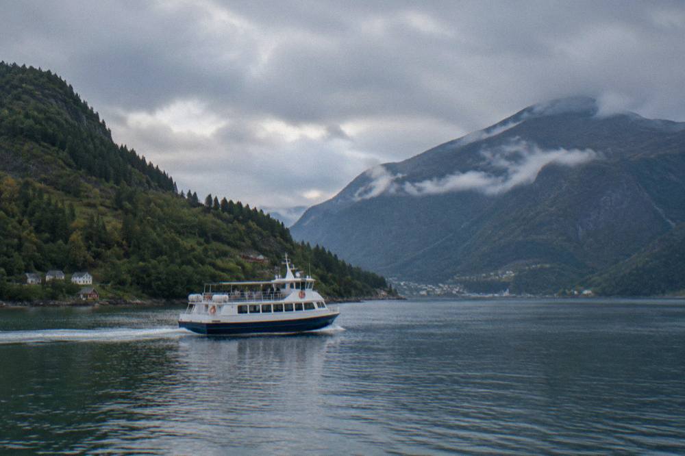 A fjord boat cruising past forested Norwegian mountainsides on a calm waterway