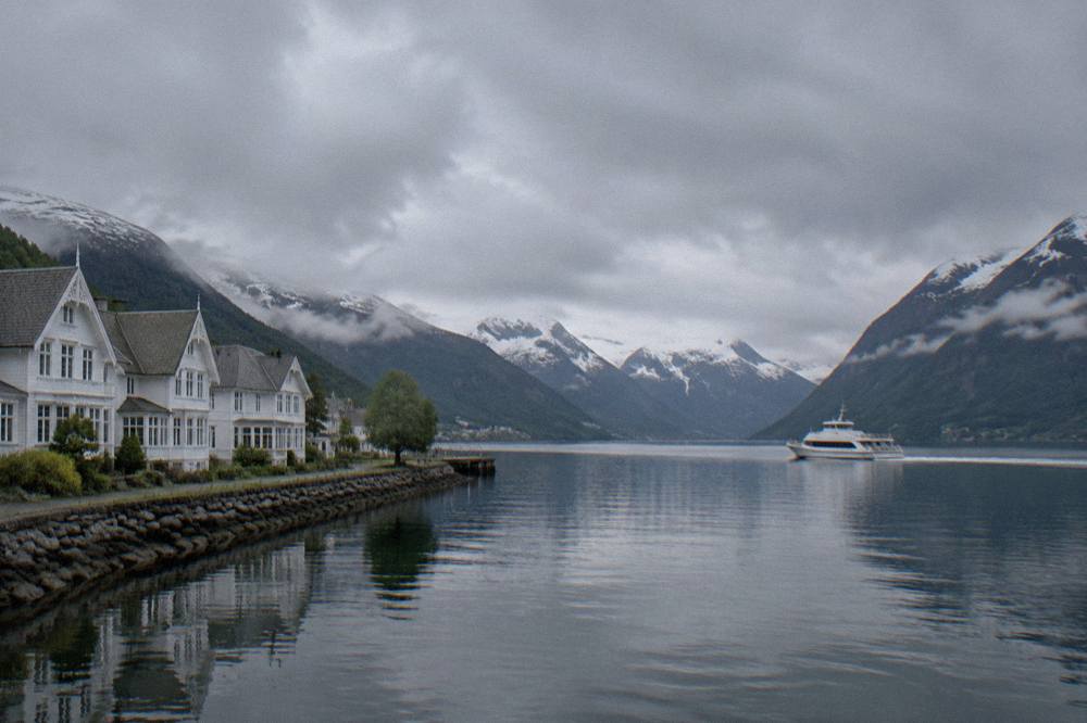 A white express boat crossing open fjord water with steep mountains on both shores under a cloudy sky