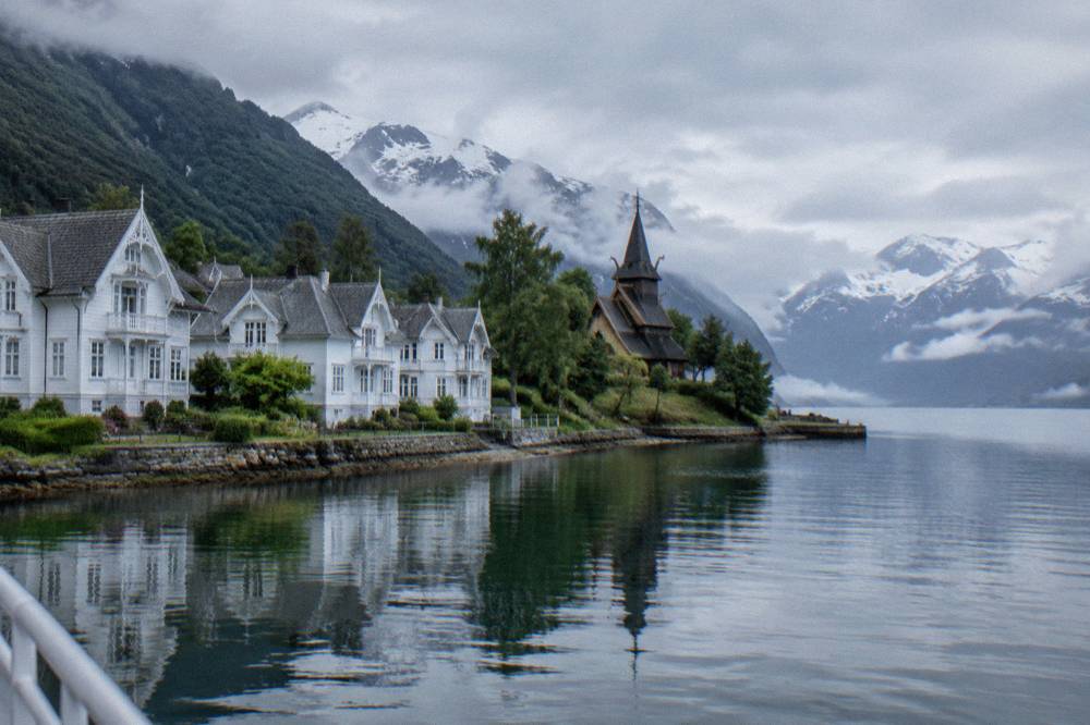 A small Norwegian church with tiered roofs and carved dragon heads standing among trees near the fjord