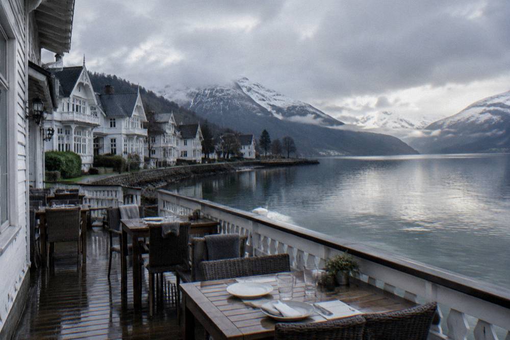 A wooden dining veranda overlooking a fjord, empty tables set for lunch with a low railing and water beyond