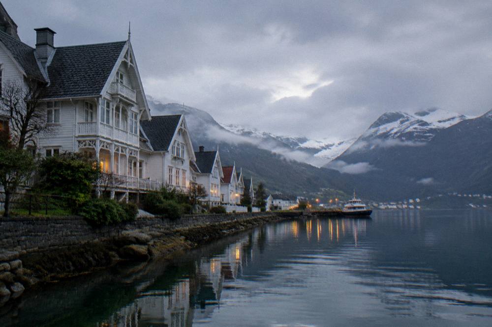A calm fjord at dusk with a village's lights reflecting on the water and dark mountains behind