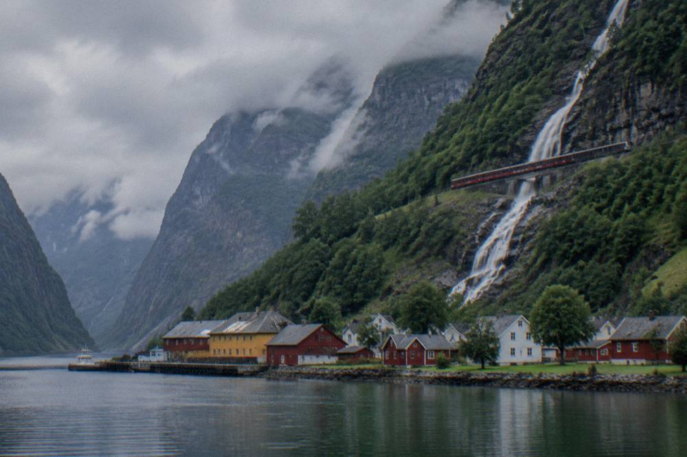 A red Flåm Railway train climbing beside a waterfall on the steep mountainside above the village