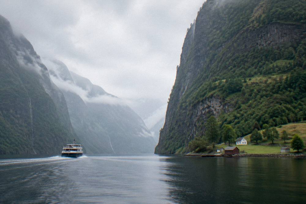 A sightseeing boat cruising between steep cliffs and a small farm on the shore of the narrow Nærøyfjord