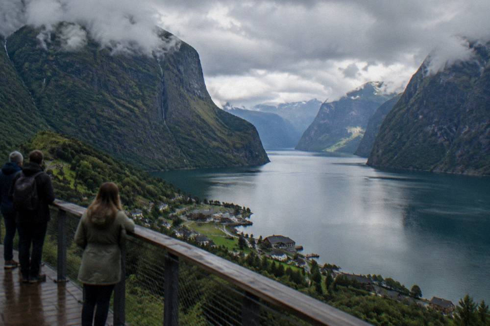 Travellers on a wooden viewpoint platform looking out over the blue water of the Aurlandsfjord between steep valley walls