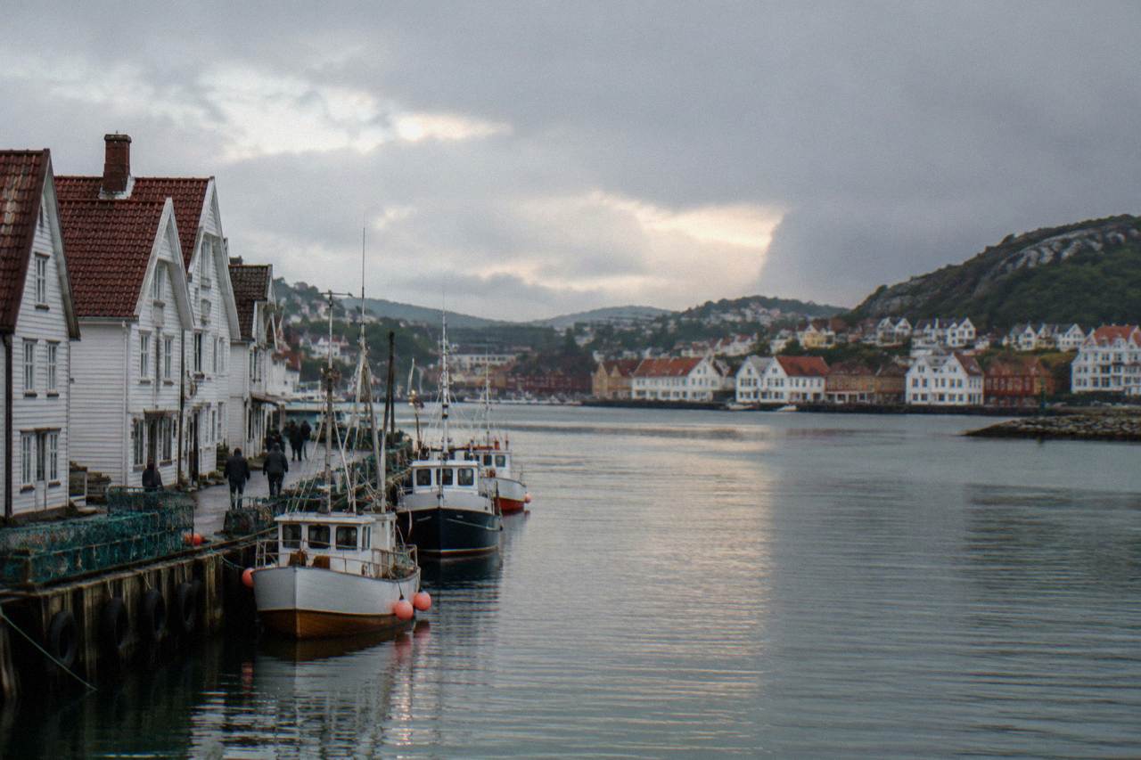 Two Norwegian coastal cities seen across calm water with wooden buildings along the shoreline and green hills rising behind them