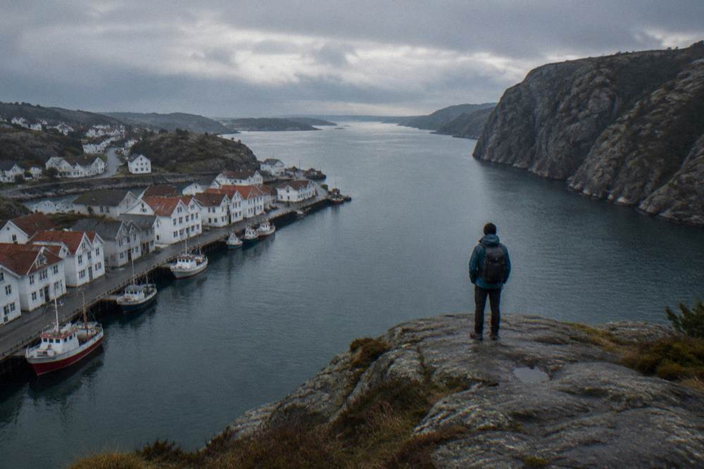 A hiker standing on a flat cliff plateau looking down over a narrow fjord with steep rock walls