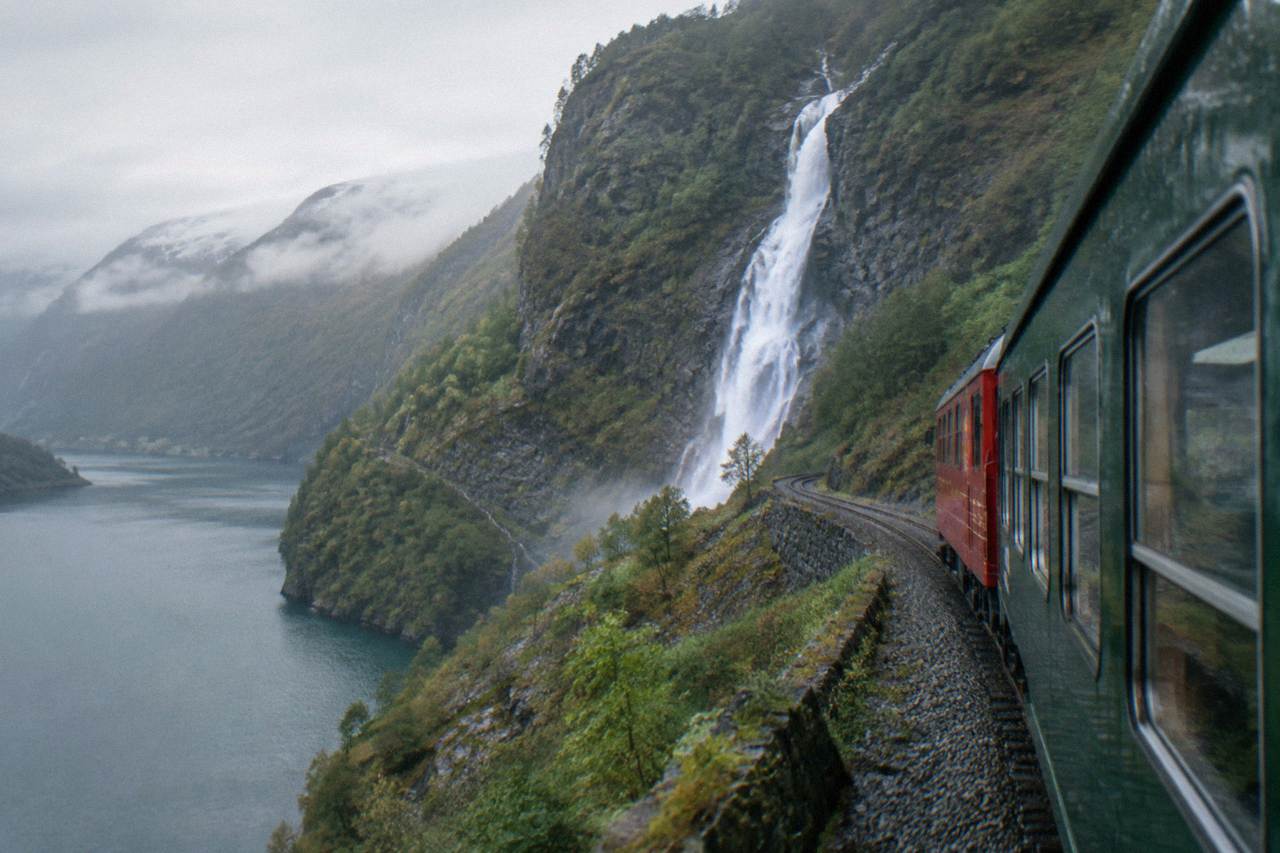 A red and green railway carriage climbing a steep green valley with a waterfall in the distance