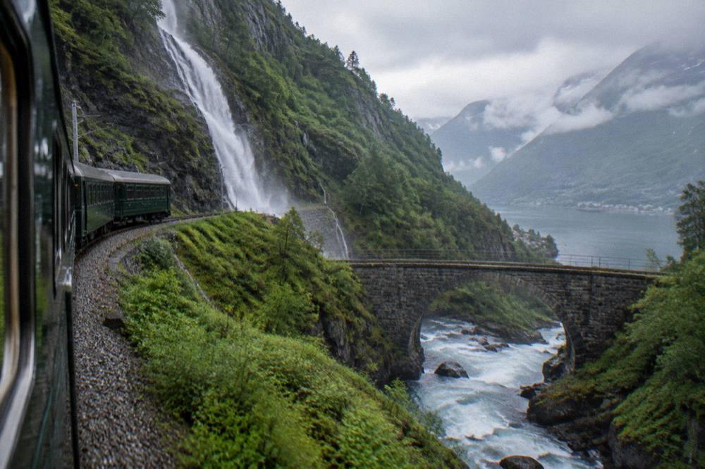 A mountain train crossing a stone bridge over a rushing river in a deep valley