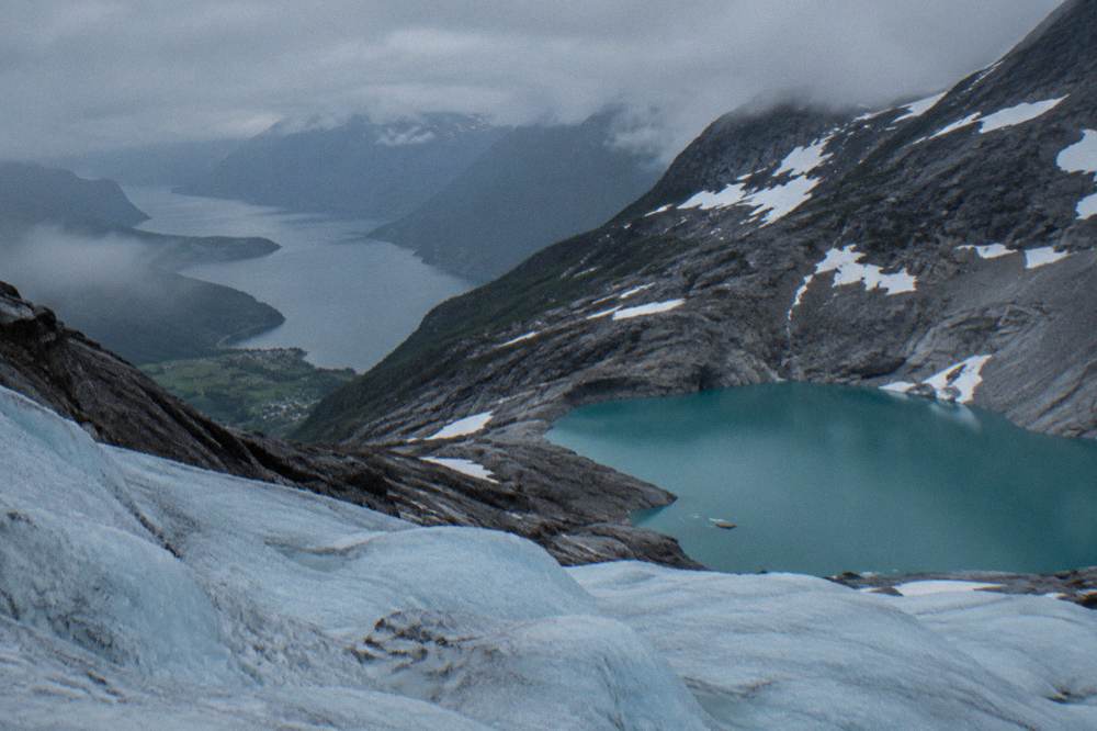 A turquoise mountain lake below a glacier with bare grey rock slopes and patches of snow around it