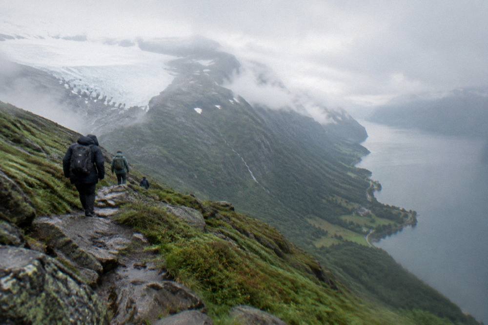 A steep mountain trail above a fjord with green slopes dropping to grey water far below