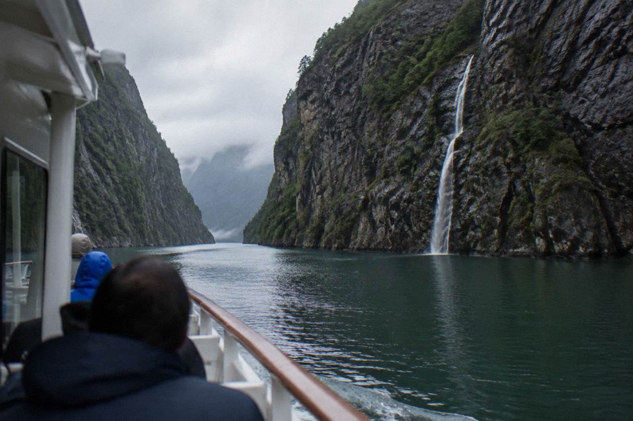 A passenger boat cruising a narrow fjord strait between steep dark cliffs in western Norway