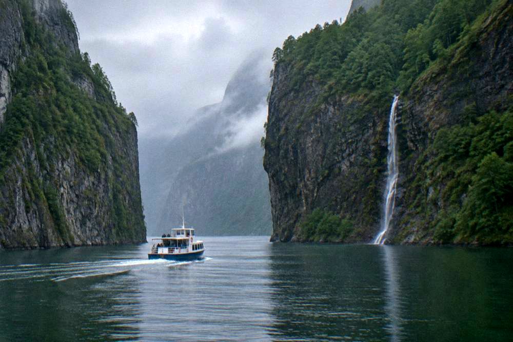 Steep forested cliffs rising from calm fjord water with a small waterfall on one face