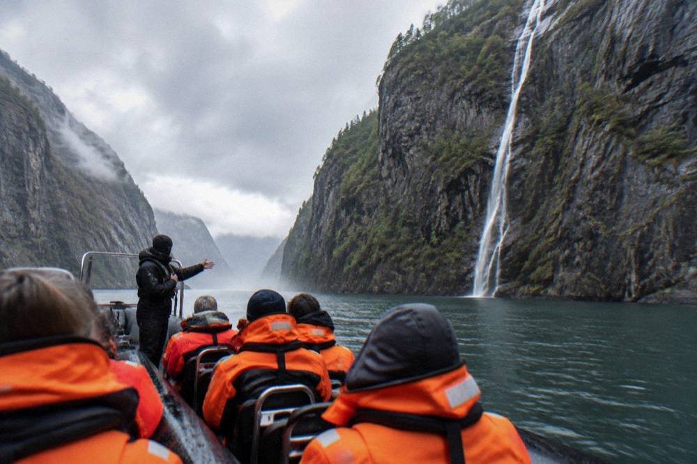 A crew member briefing passengers in orange survival suits on the deck of a RIB boat