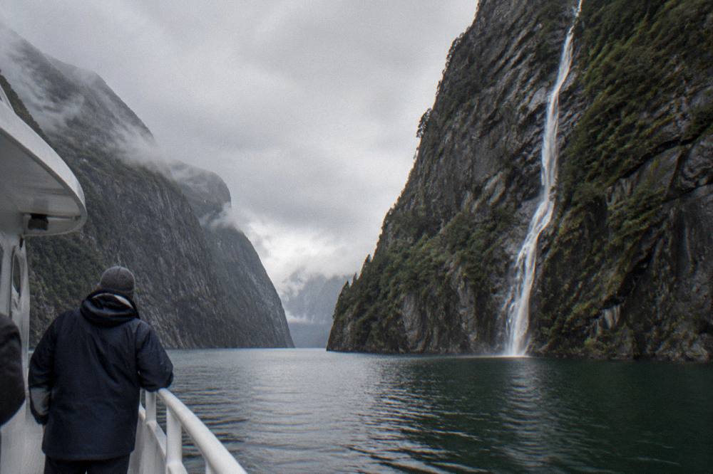A passenger in a waterproof jacket and hat standing at a boat rail looking at fjord cliffs