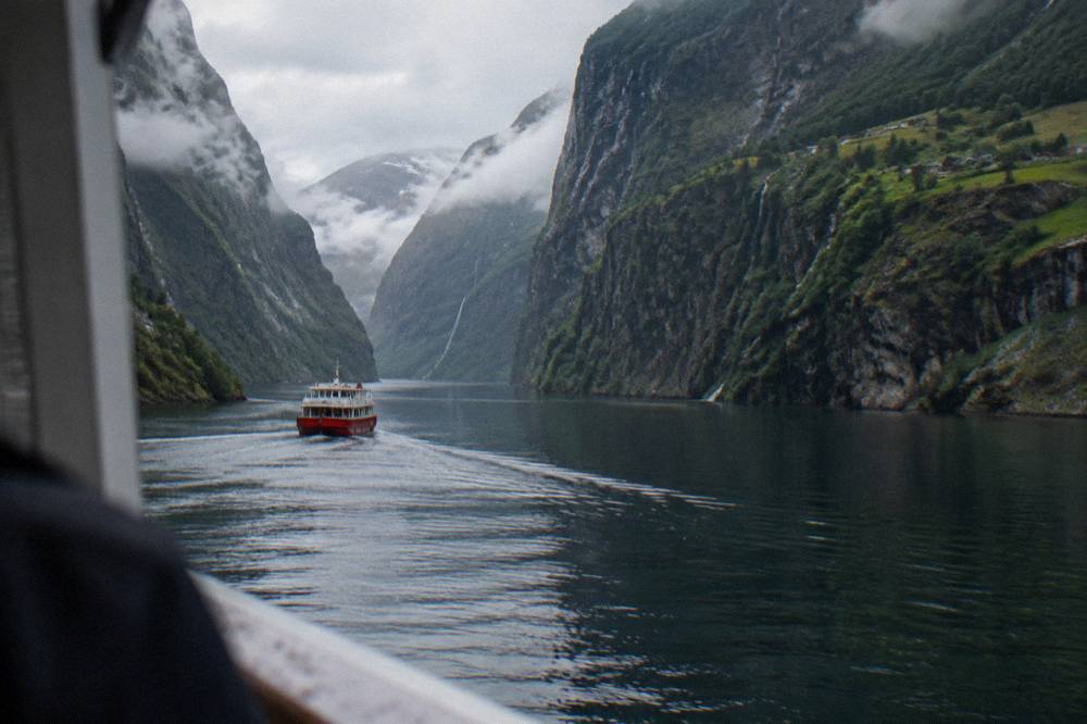 A red passenger boat travelling along a narrow fjord between dark rock walls