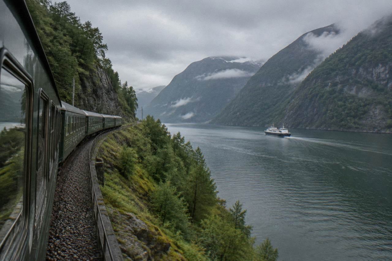 A scenic railway train winding along a fjord through green Norwegian mountains