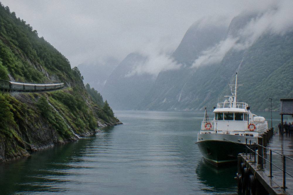 A passenger boat docked at a small fjord pier with steep forested mountains rising behind it