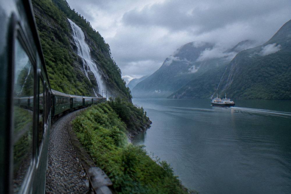 A mountain railway track curving beside a tall waterfall in a green Norwegian valley