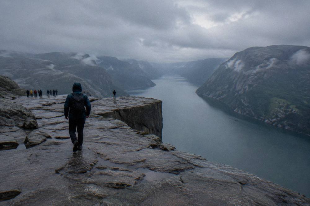 A hiker in rain gear standing on a bare rocky plateau looking out over a fjord