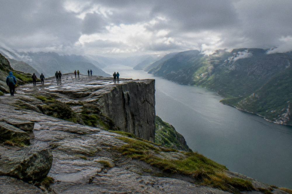 A rocky cliff edge overlooking a long narrow fjord with steep green mountainsides fading into haze