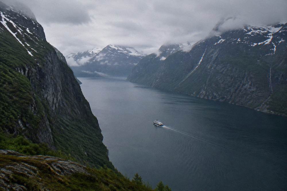 A white boat seen from above on dark fjord water winding between two near-vertical rock faces