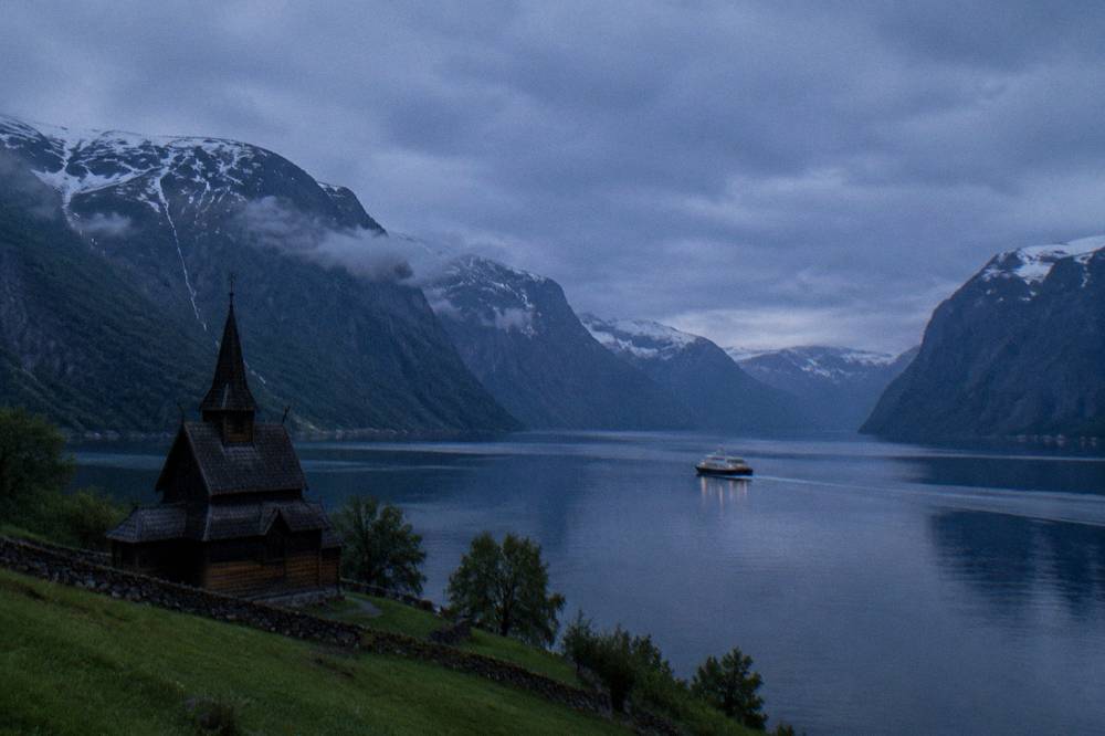A wooden stave church beside a calm fjord with mountains reflected in still water