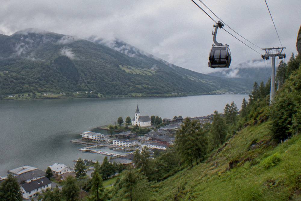 A gondola cabin ascending a steep green hillside above a lake and a small town