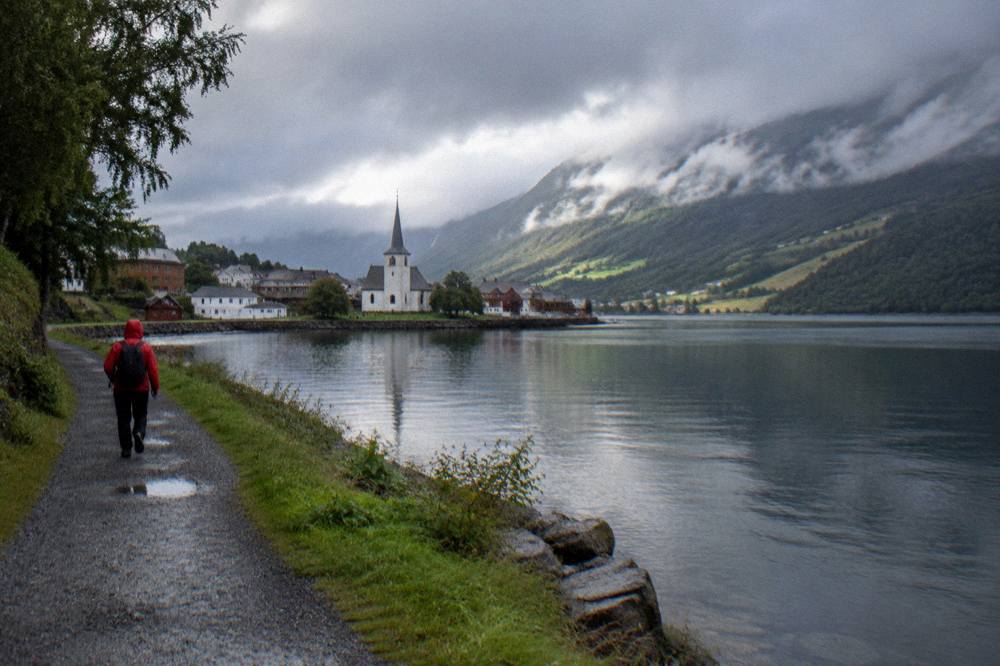 A hiker in a red rain jacket walking a gravel path beside a calm lake under low clouds