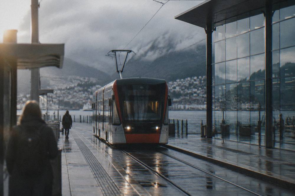 A light rail tram arriving at an outdoor station platform beside a glass airport facade