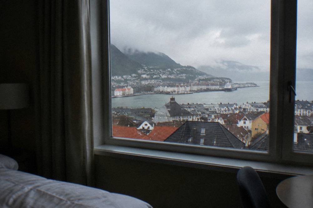 A hotel room with a window overlooking illuminated rooftops of a coastal town at dusk