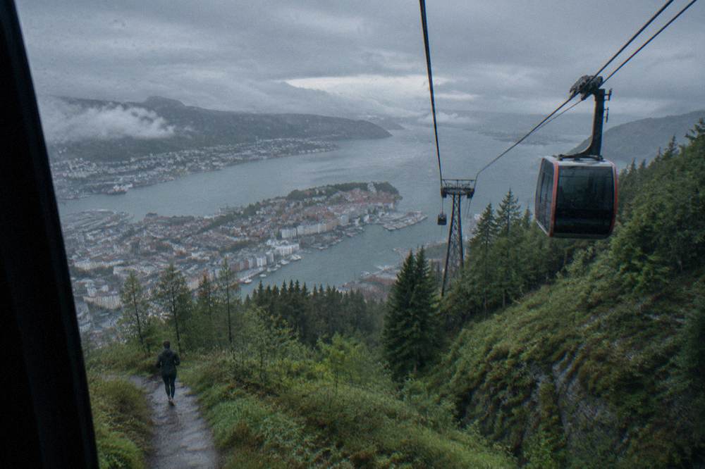 A cable car cabin ascending a forested mountain above a coastal city under overcast skies