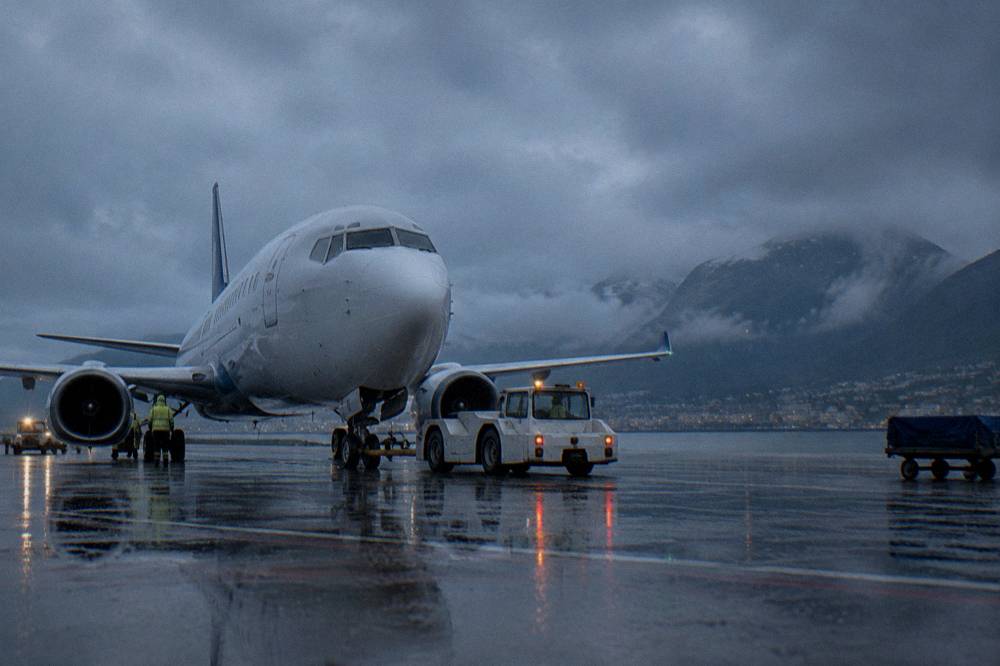 Rain falling on an airport tarmac with a parked aircraft and ground crew vehicles under grey skies