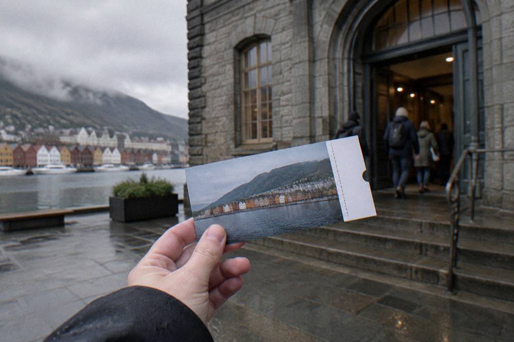 A hand holding a paper sightseeing pass in front of a museum entrance in Bergen