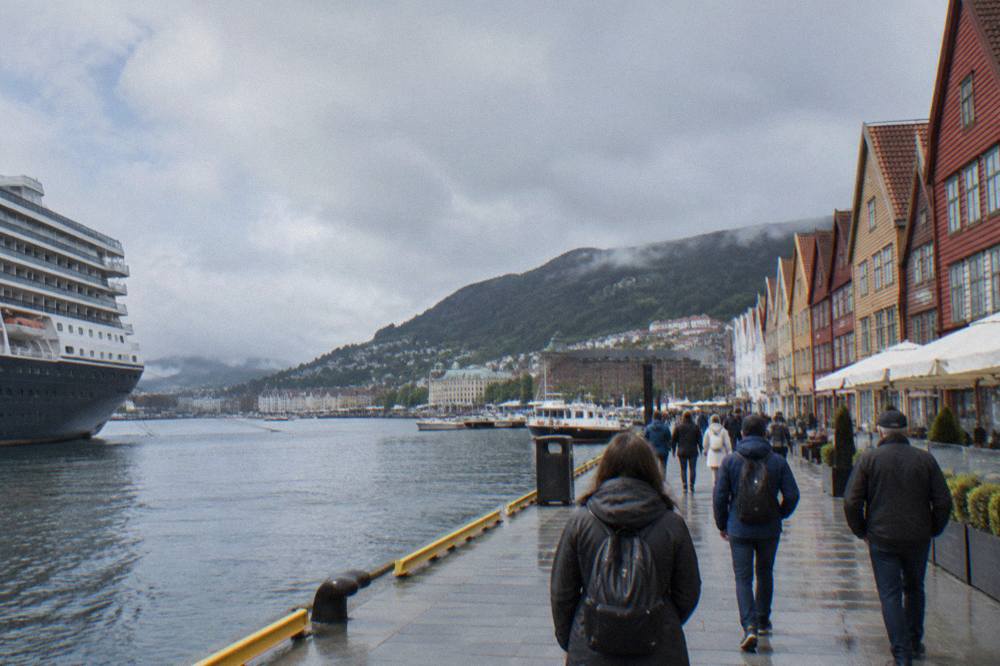 Passengers walking along a waterfront promenade from a cruise ship toward old wooden harbour buildings