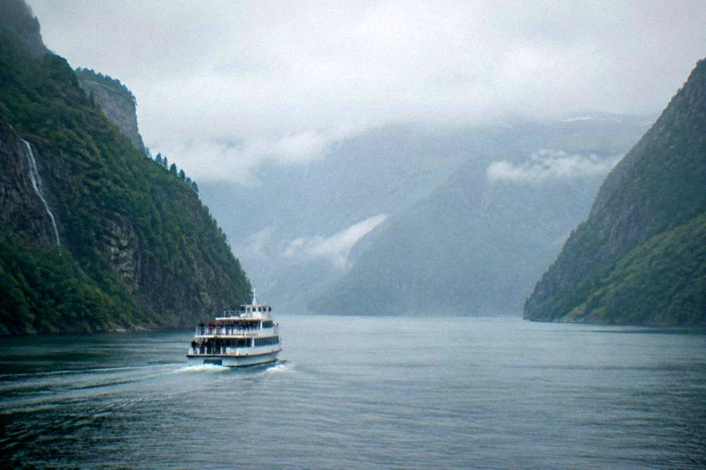 A tour boat cruising on a narrow fjord between steep green cliffs in western Norway