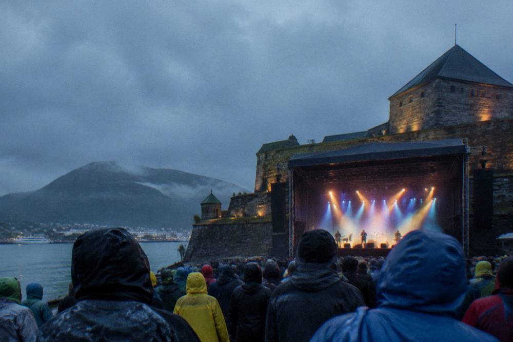 Concertgoers with rain jackets standing in front of a lit stage at a fortress music festival in Bergen, Norway