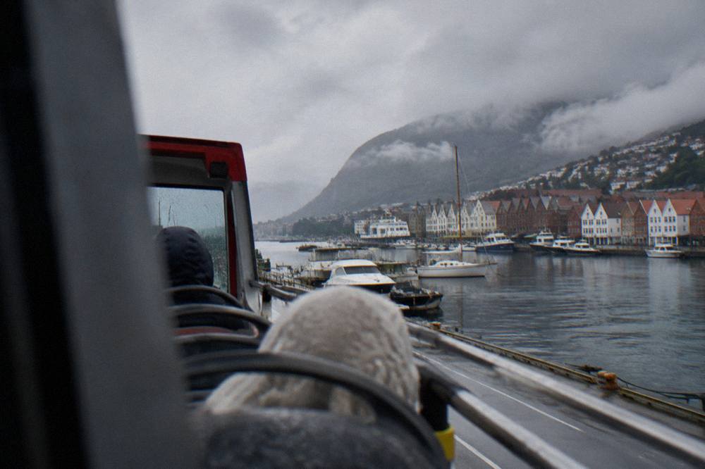 A covered open-top sightseeing bus driving along a waterfront road with small boats moored in the harbour