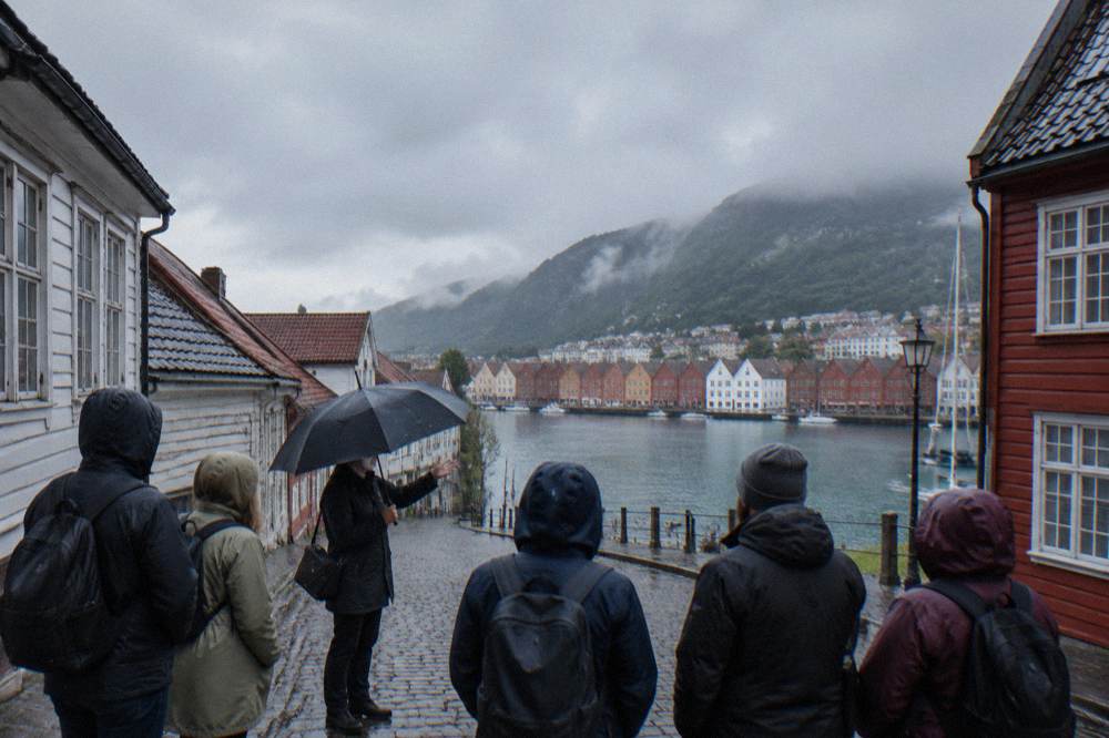 A small group of tourists with an umbrella-wielding guide listening to a talk on a cobbled street between old timber buildings