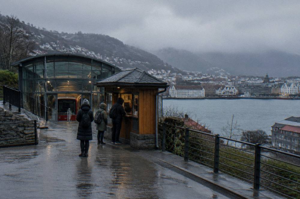 Travellers buying tickets at a small wooden kiosk beside a funicular station entrance on a Bergen street