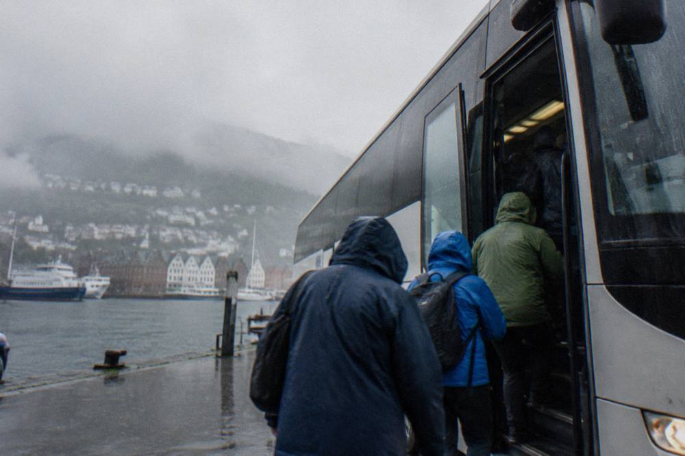 Passengers in rain jackets boarding a sightseeing bus while rain falls on the harbour street around them
