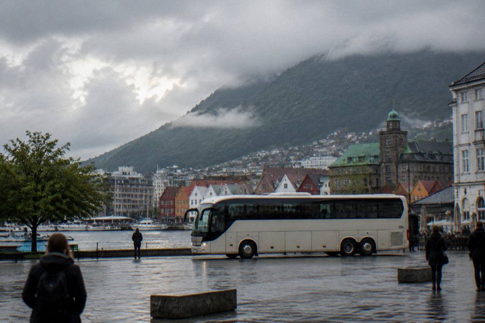 A sightseeing bus crossing an open square with mountain ridges rising behind the rooftops of Bergen in the background