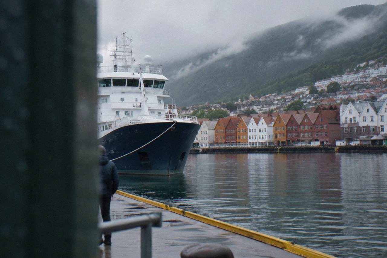 A coastal expedition ship moored at a dock in Bergen with colourful wooden buildings along the waterfront behind it