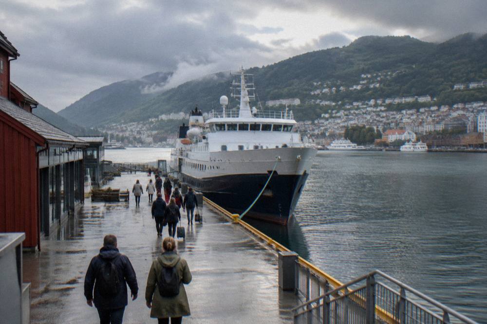 Passengers walking along a quay toward a moored coastal ship with mountains rising behind a Norwegian harbour town