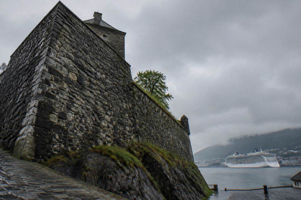 A historic stone fortress wall beside the harbour in Bergen with a cruise ship at anchor in the background
