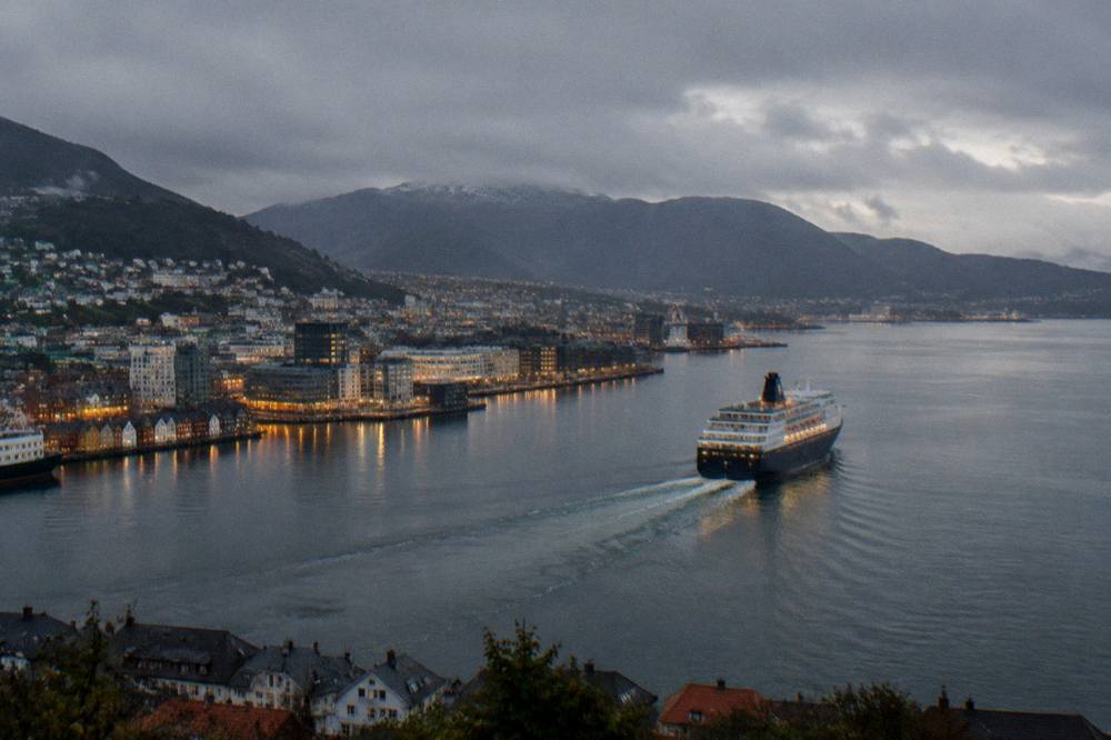 A coastal ship sailing away from Bergen at dusk with the illuminated city and mountains behind it