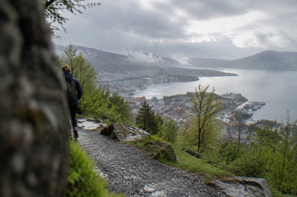 A hiker on a gravel trail above Bergen with the city and fjord visible below in spring light
