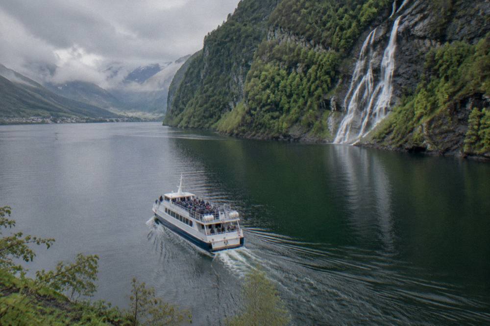 A tour boat on a calm fjord arm near Bergen with steep green cliffs and a waterfall in spring