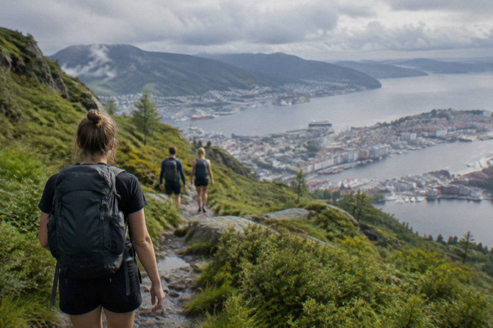 Hikers walking a green mountain trail above Bergen with the city and fjord visible below